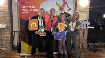 Four white presenting women pose in front of a Mission Soil banner holding card board cut outs of soil ecosystem elements.