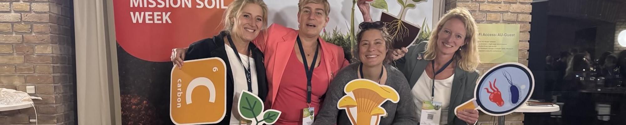Four white presenting women pose in front of a Mission Soil banner holding card board cut outs of soil ecosystem elements.