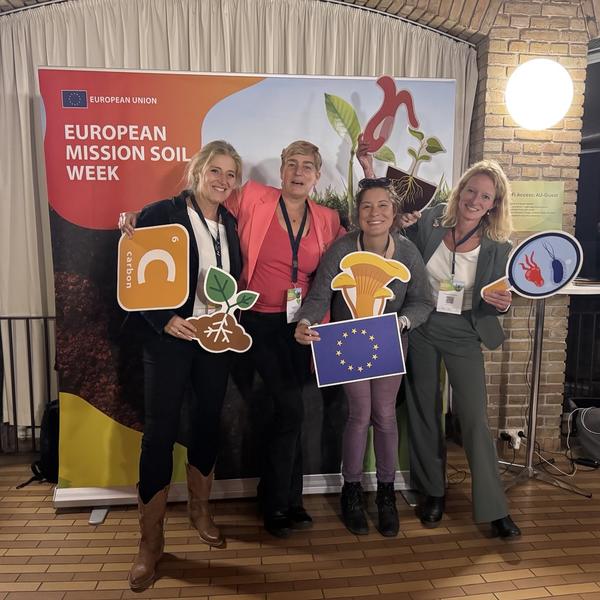 Four white presenting women pose in front of a Mission Soil banner holding card board cut outs of soil ecosystem elements.
